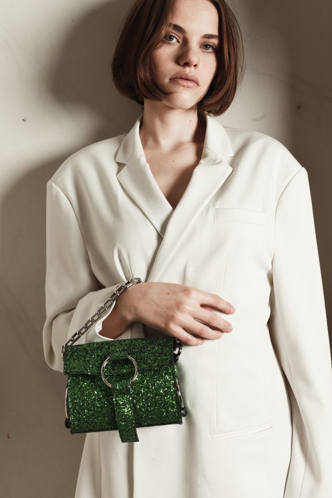 Woman in white blazer holding a sparkling green glitter chain-strap handbag against a neutral wall.