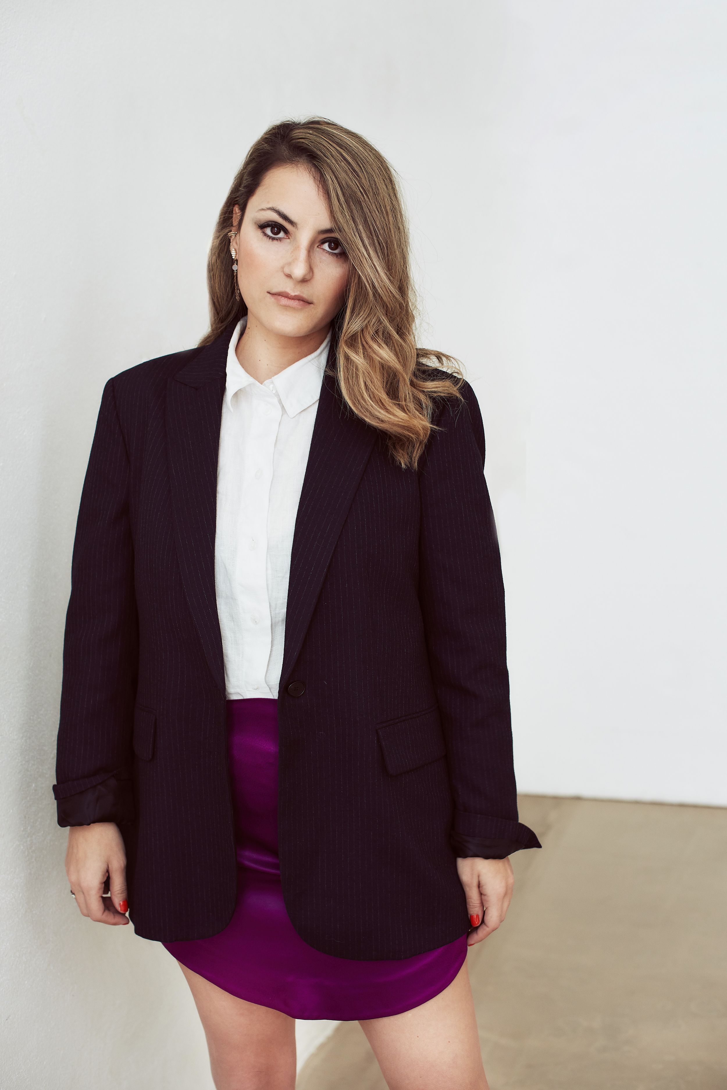 Woman in a black blazer, white shirt, and purple mini skirt poses confidently against a white wall.