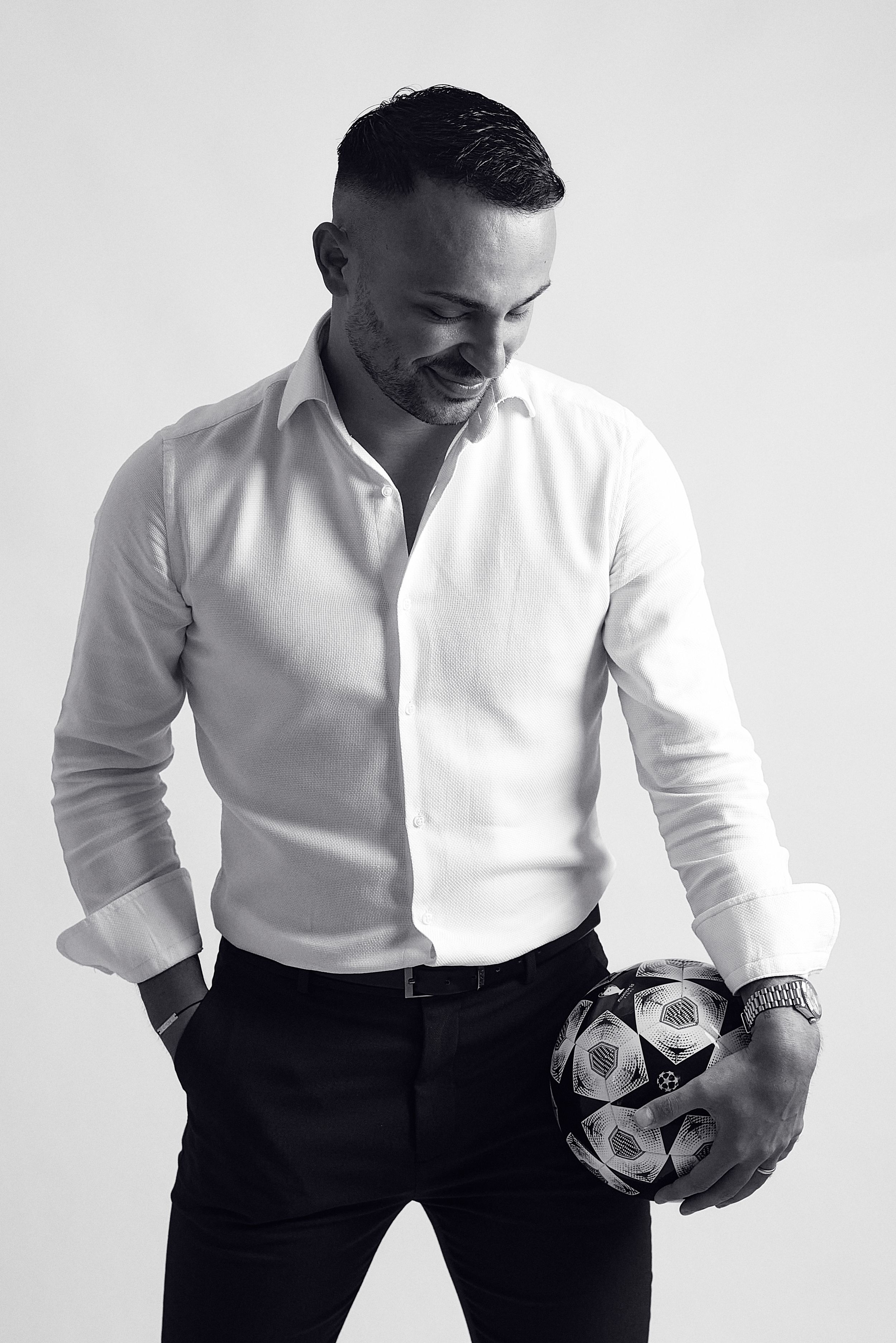 Stylish man in white shirt holding a soccer ball looking down in a black and white studio portrait.