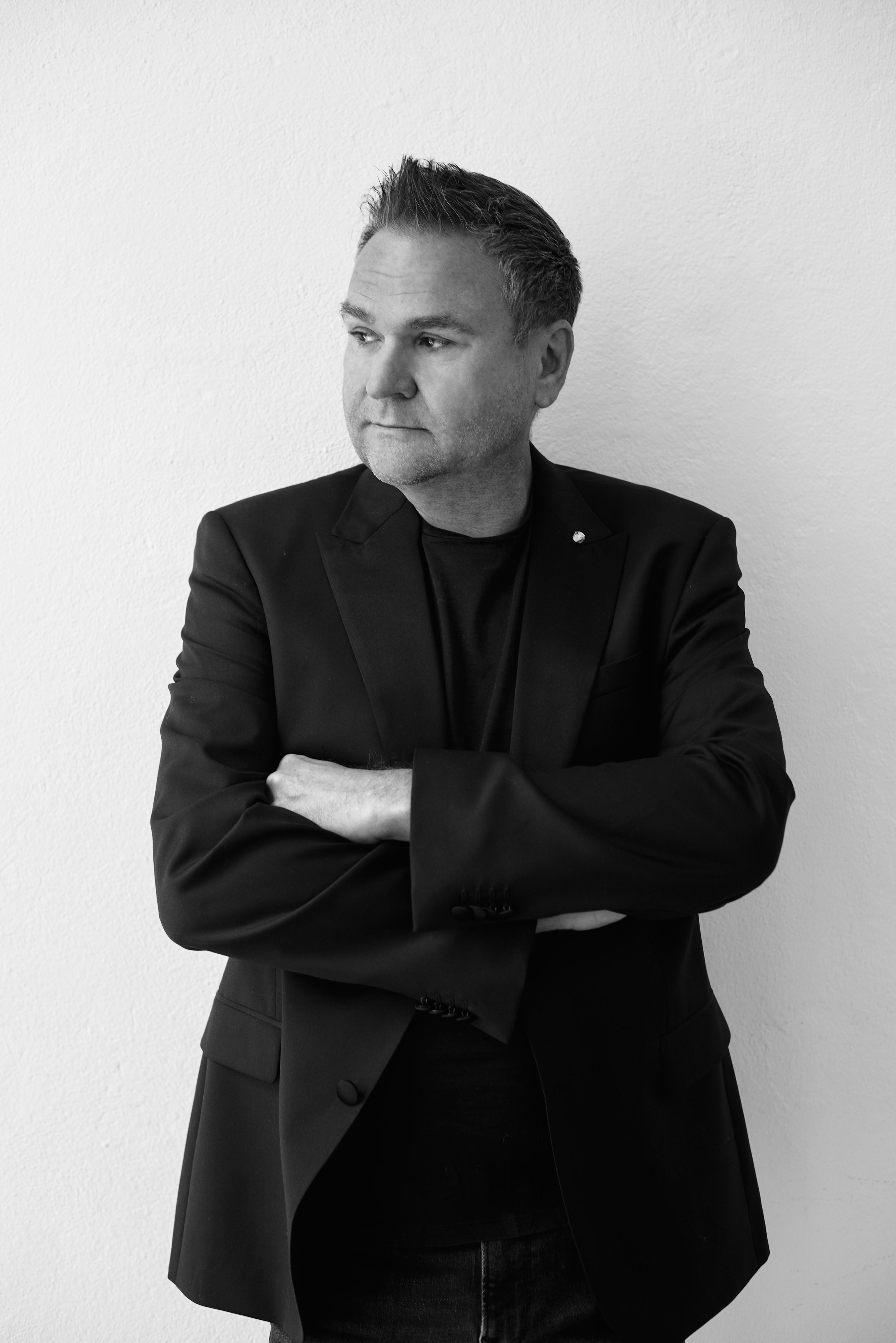 Man in black blazer with arms crossed leaning against a white wall in a black and white portrait.