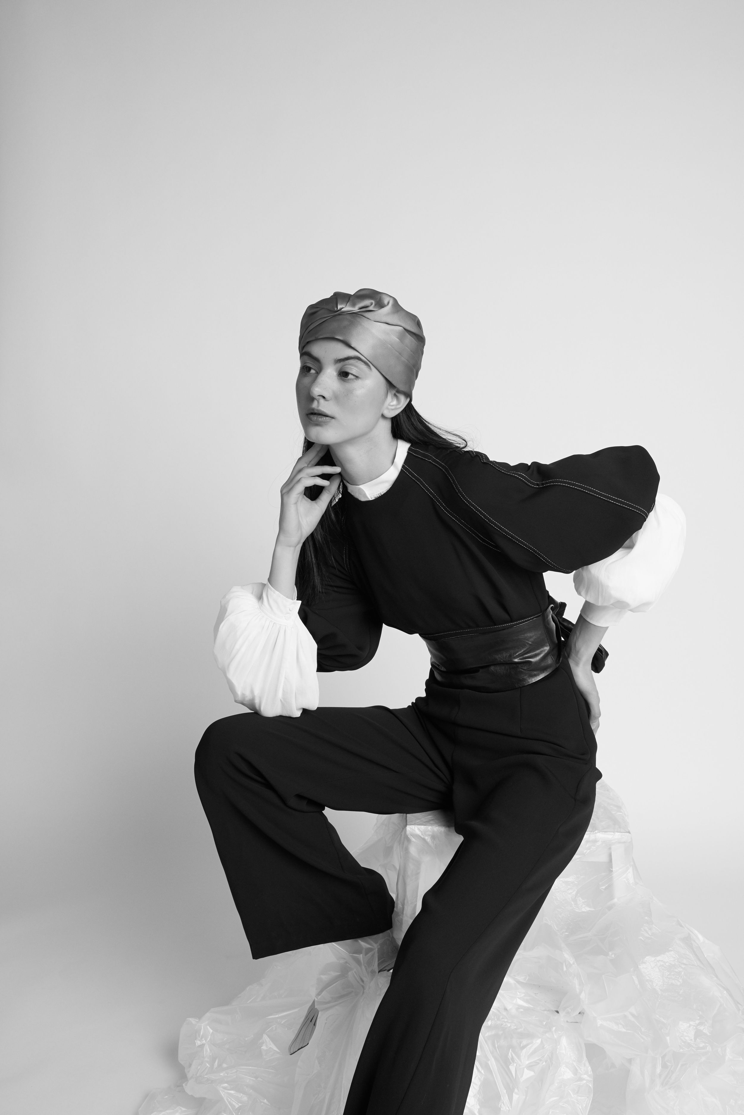 Black and white fashion portrait of a model in black outfit with leather belt and metallic cap.