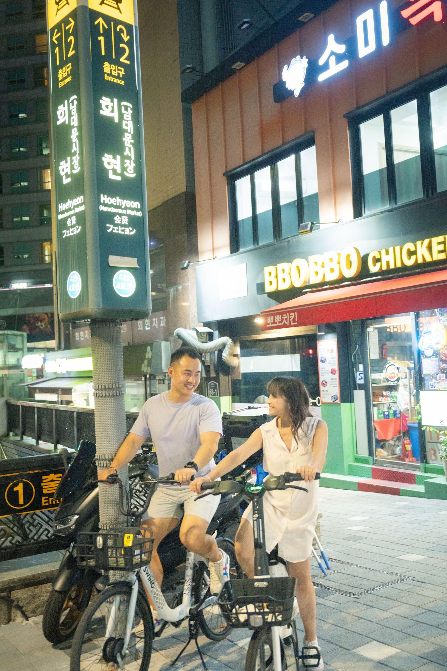 People riding bicycles on an urban street at night with illuminated Korean storefront signs and restaurants.