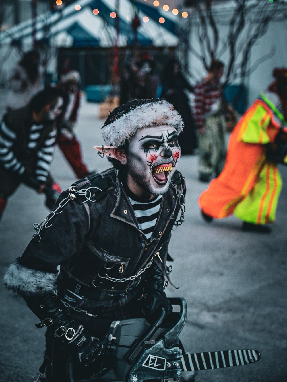 Dark atmospheric carnival scene with performers in spooky monochrome costumes and face paint.
