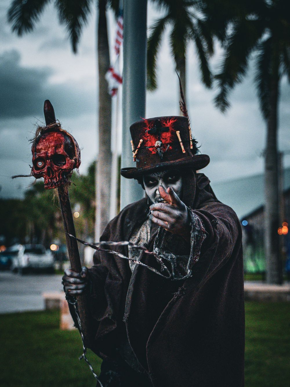 Sinister figure in dark coat and skull mask poses with prop weapons against palm tree backdrop.