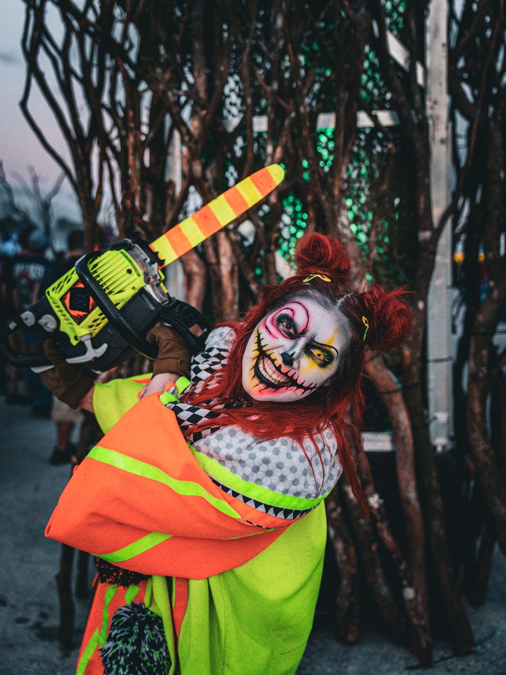 Creepy clown with chainsaw wearing neon safety gear poses menacingly in front of dark trees with green lights.