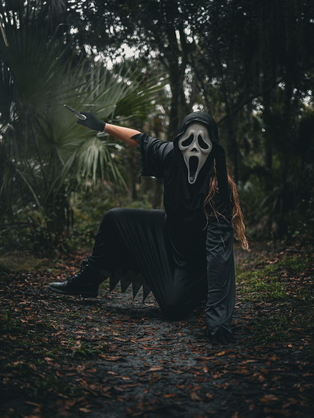 Person in ghost mask and dark clothing crouches on leaf-covered forest ground.