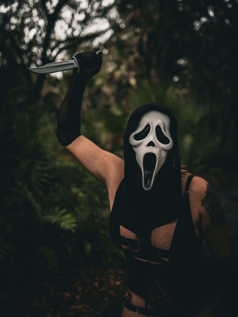 Figure wearing iconic ghost mask poses dramatically in dark wooded setting.