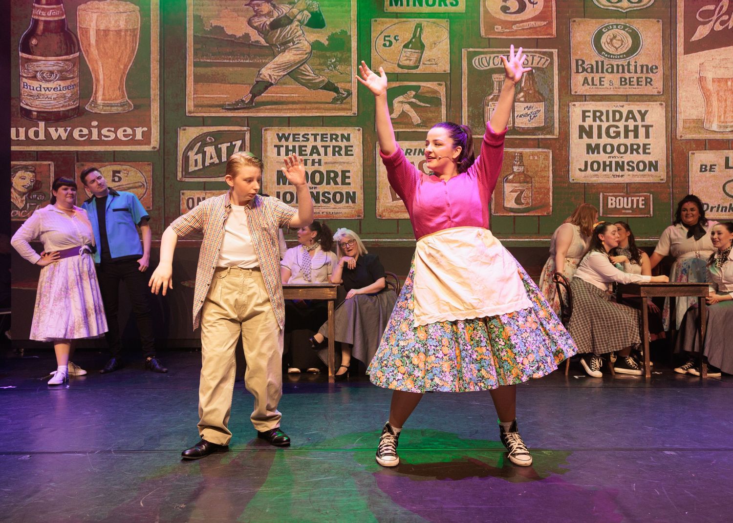 Theater stage performers dance in 1950s-style costumes against a backdrop of vintage advertising signs.