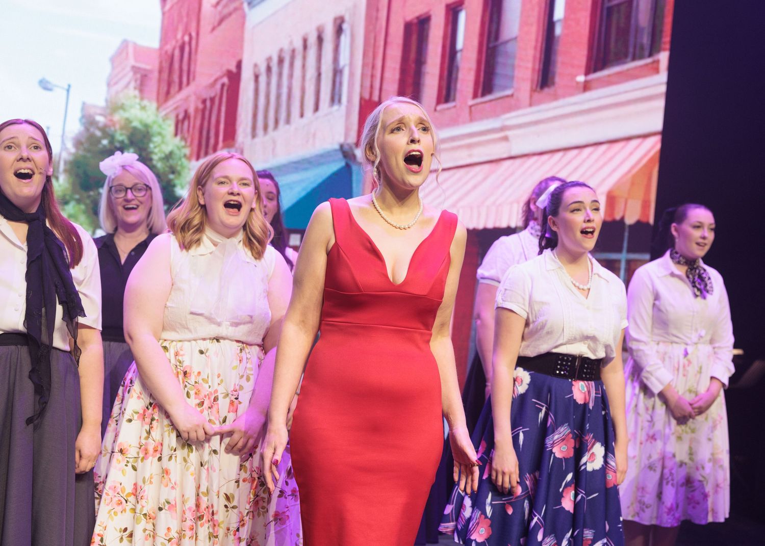 Performers in colorful vintage dresses sing together on stage during a theatrical production.