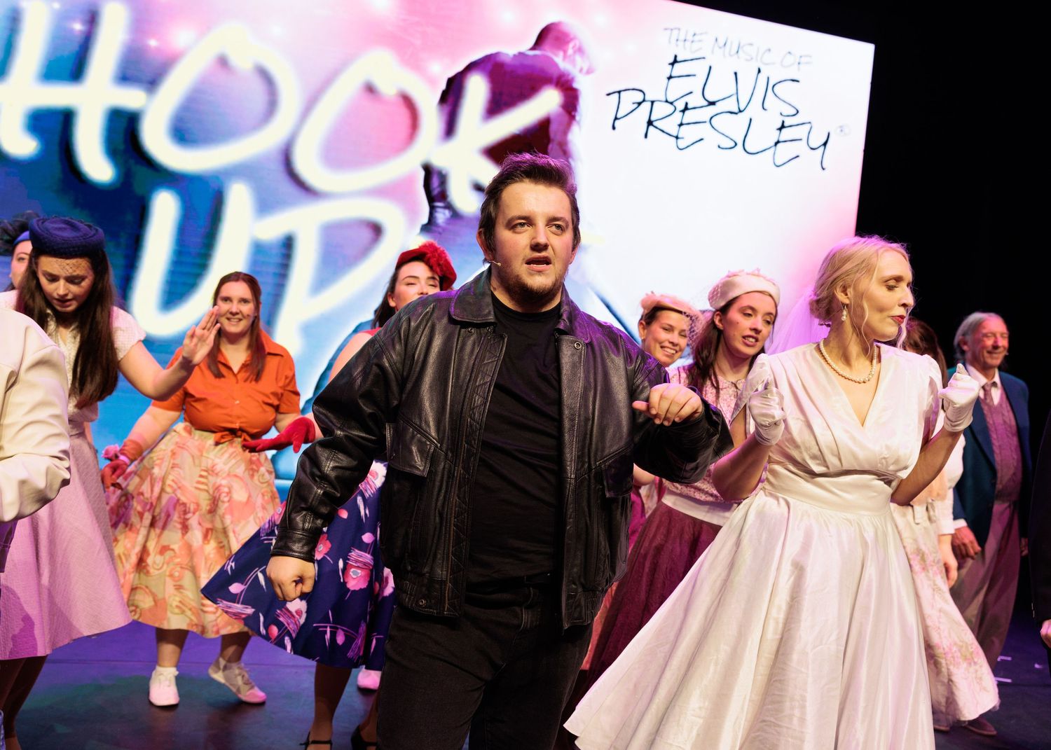 Theater performers in retro 1950s outfits dance during an energetic musical number on stage.