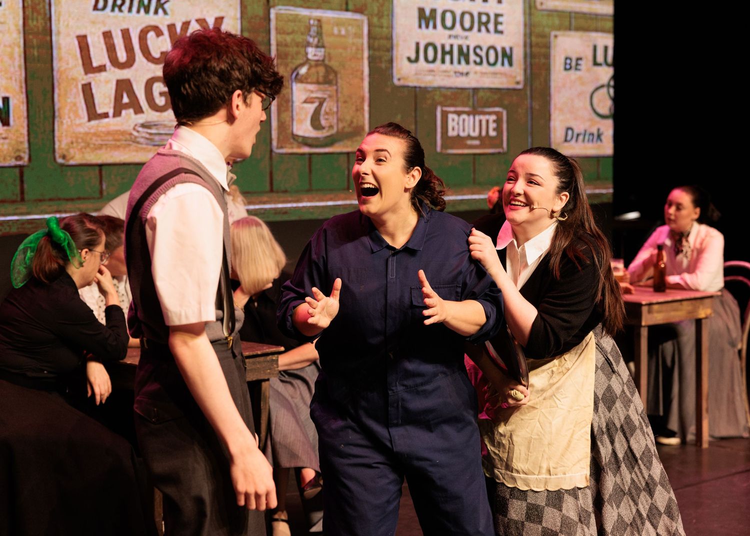 Theater performers act in an energetic scene on stage with vintage signs and decor visible in the background.