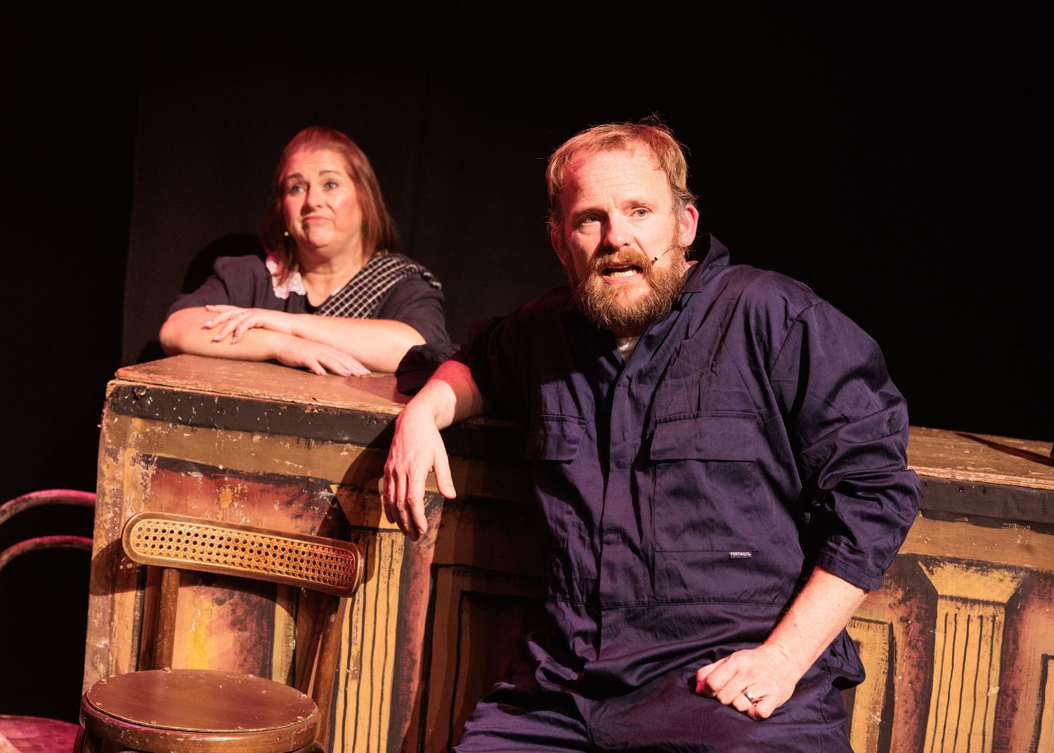 Two performers sit casually on vintage wooden theater chairs on a dark stage.