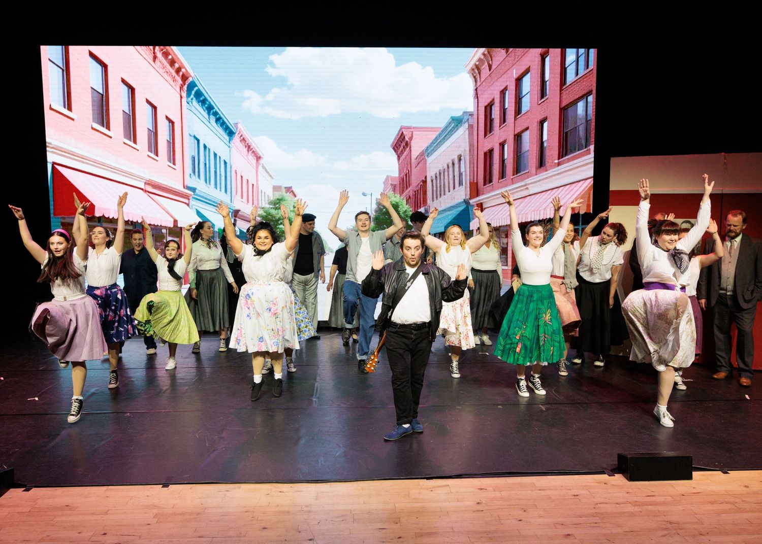 Performers in colorful costumes dance energetically across a theater stage during a lively musical production.