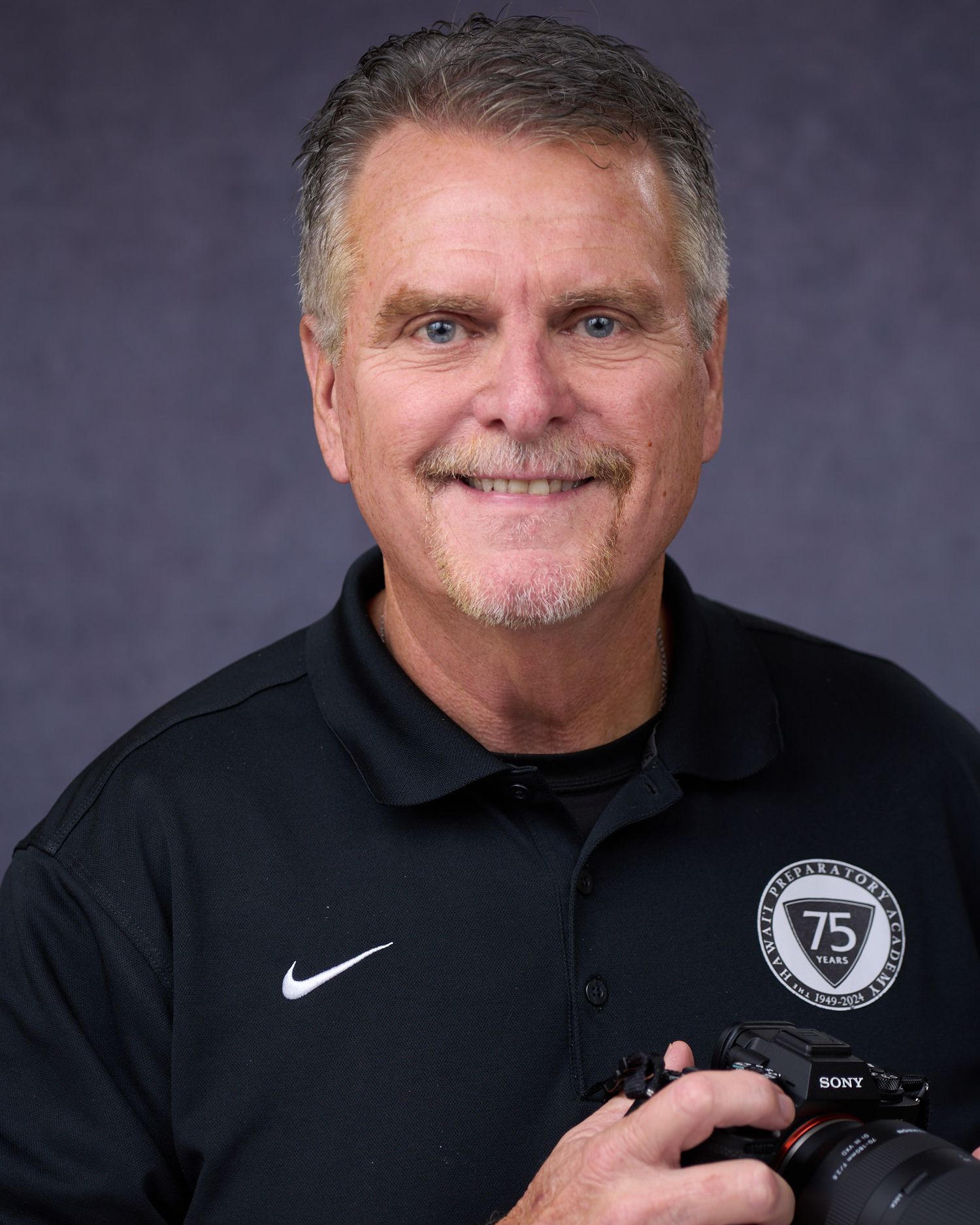 Professional male photographer smiling, holding a Sony camera, wearing a black Nike polo with a 75th anniversary badge.