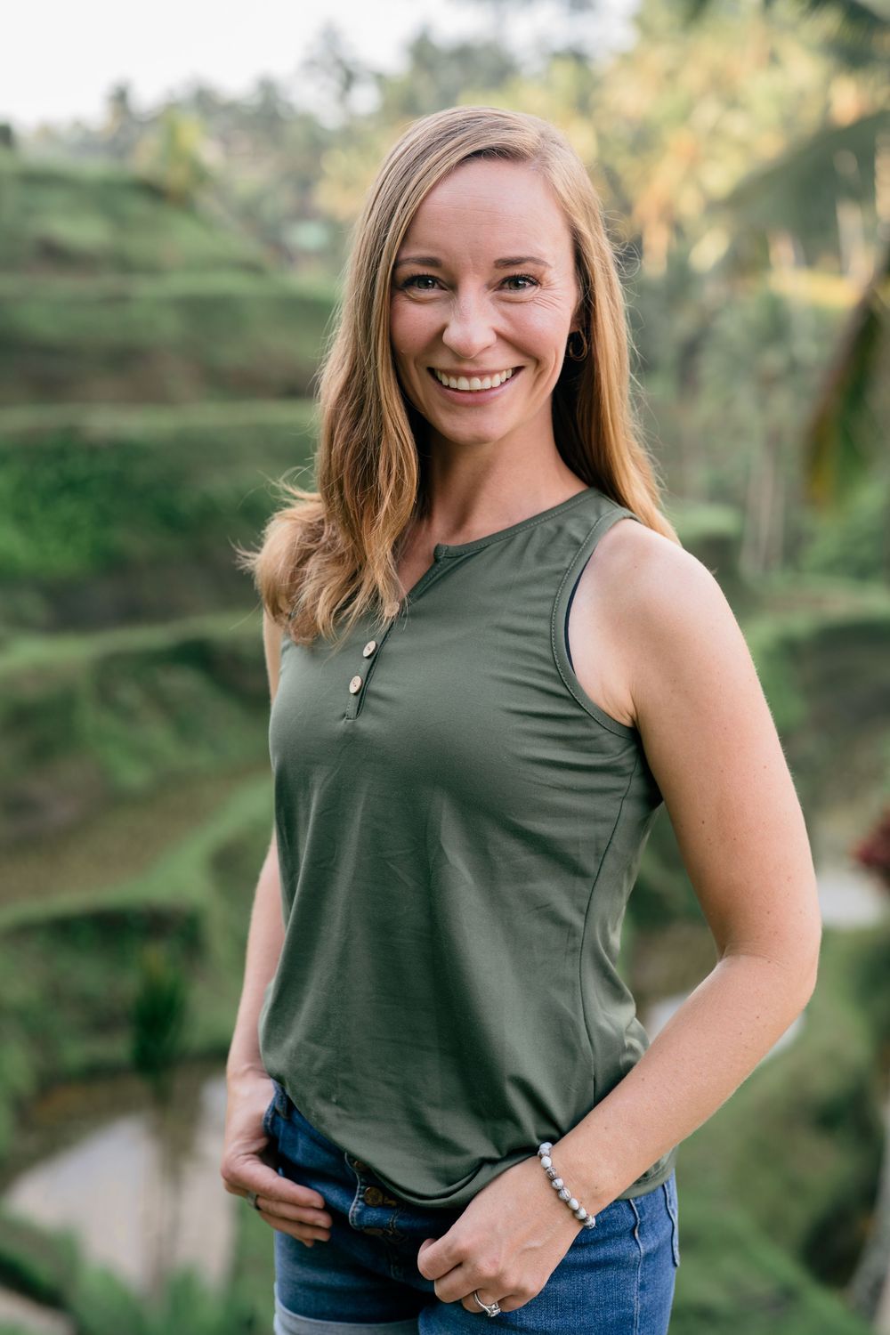 Woman in olive green tank top and jeans poses outdoors against lush green background.