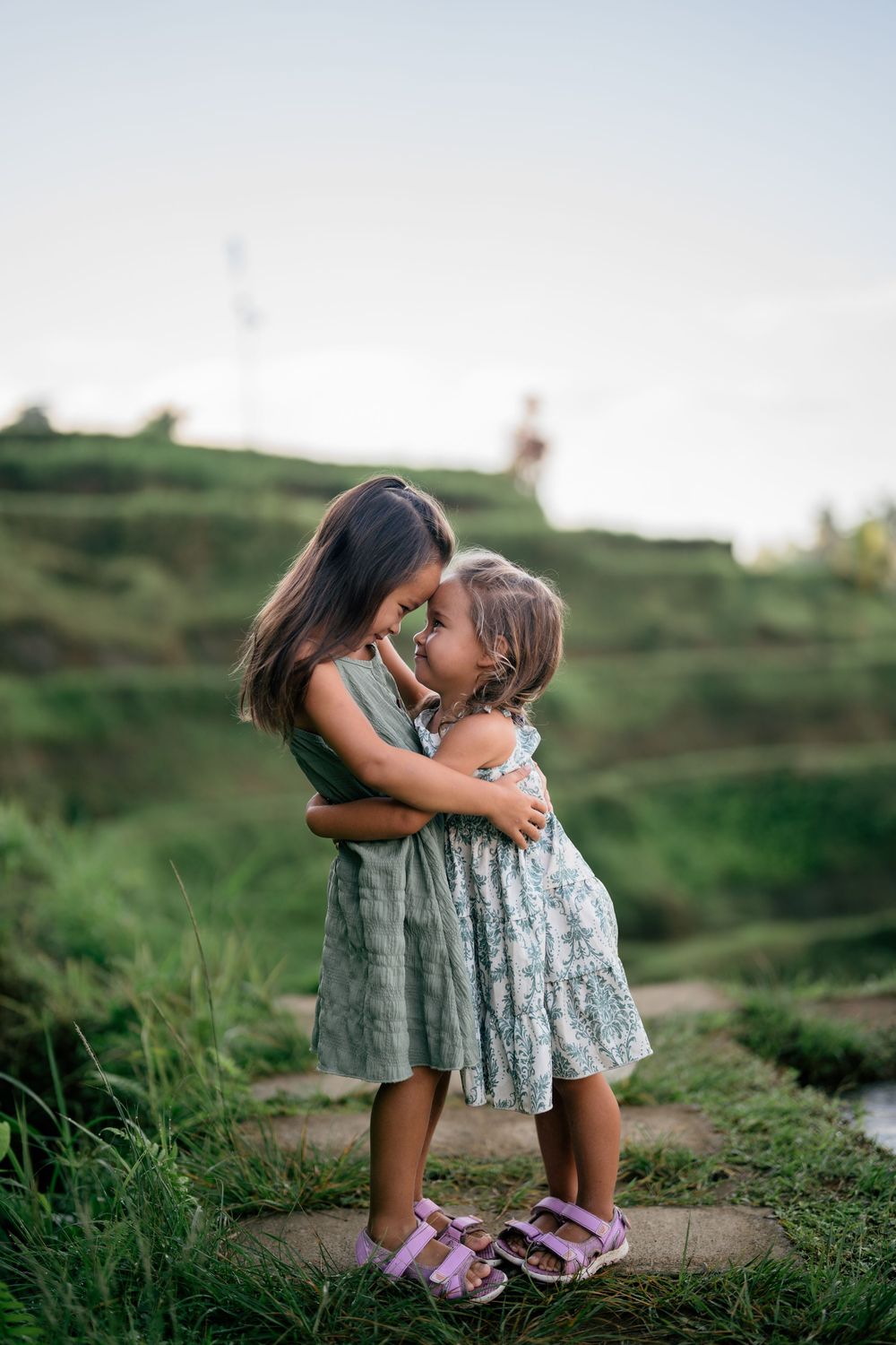 Two children in summer dresses embrace on a scenic hillside path.