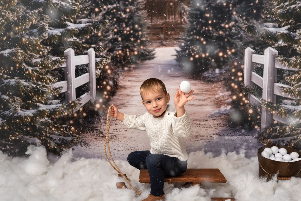 young boy sitting on the sleigh, throwing a snowball at the viewer, christmas trees, snow, white jumper, snowballs