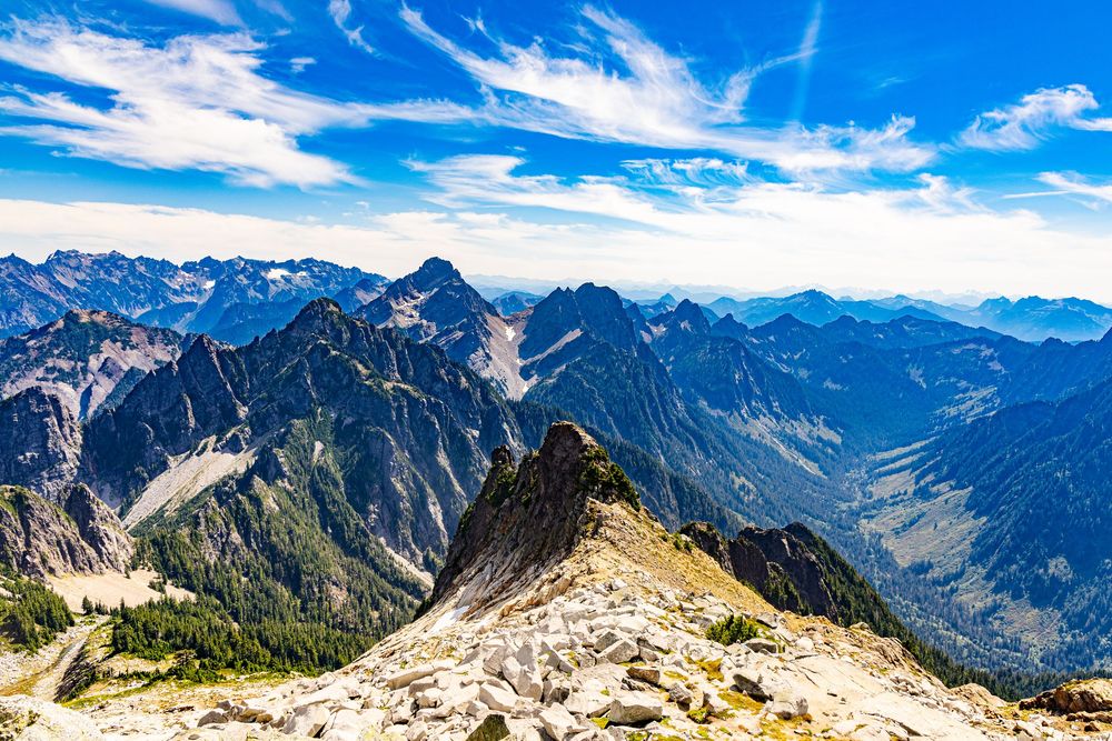 Panoramic Mountain summit view with distant peaks, rocky slope and wispy clouds through the sun.
