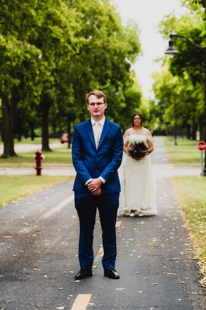 A couple in wedding attire stand at a distance on a tree-lined path during their first look moment.