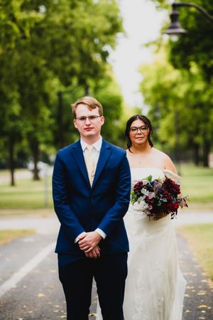 A couple poses along a tree-lined path, with the bride holding a deep red bouquet.