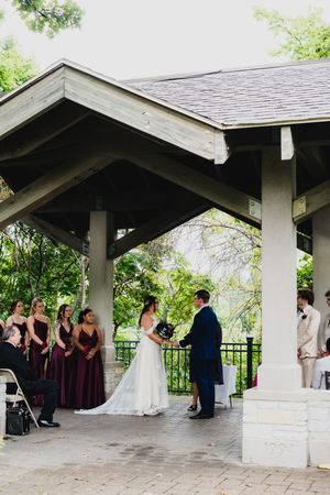 Wedding ceremony takes place under a covered outdoor pavilion with guests in burgundy dresses.