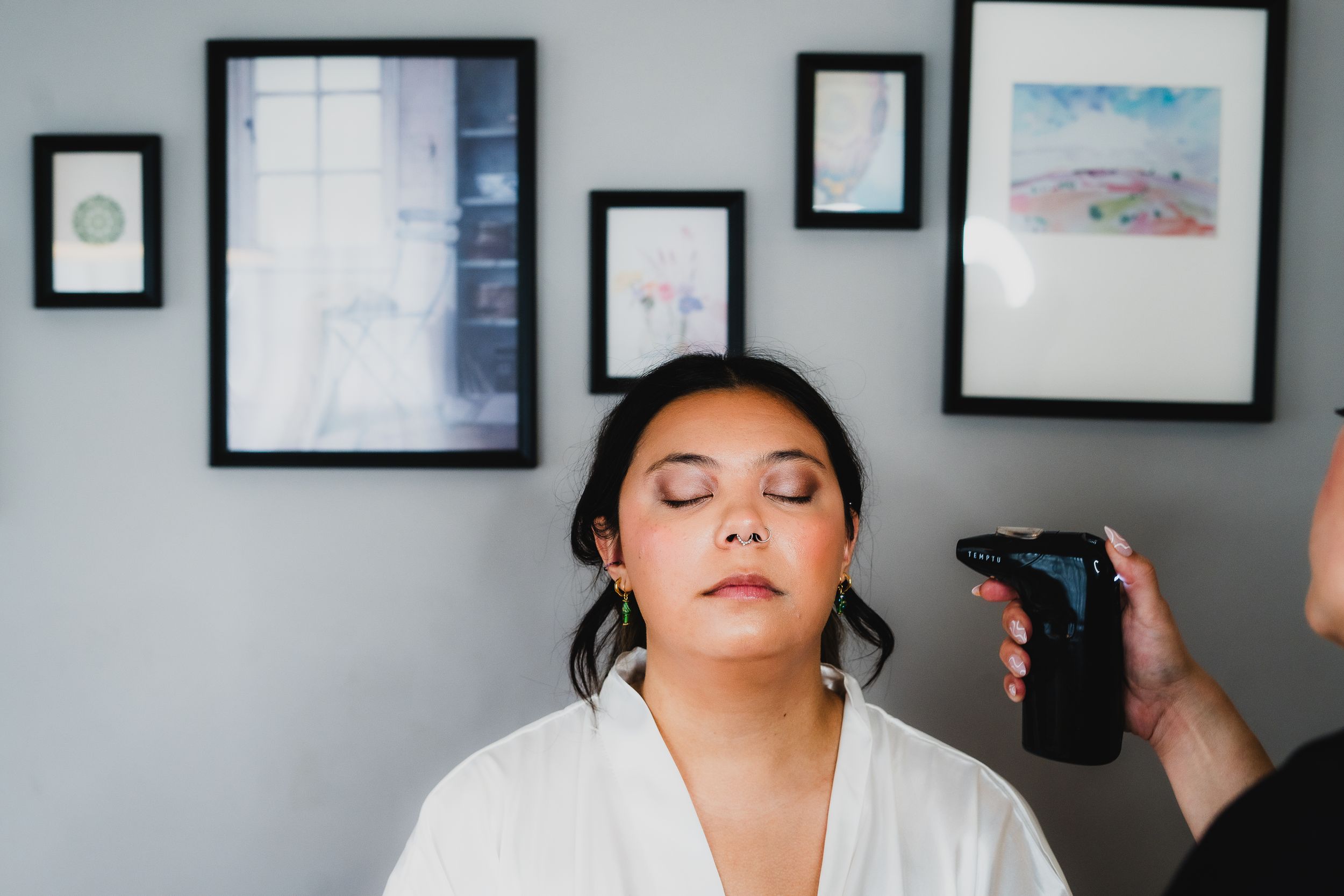 A person in a white robe getting their hair styled while sitting in front of a gallery wall with framed artwork.
