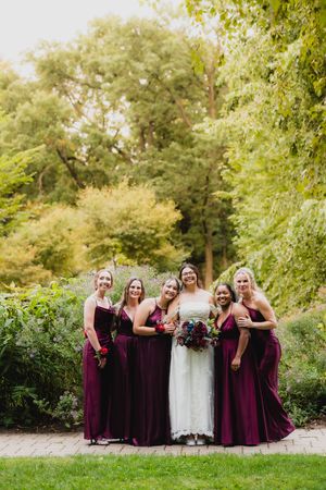 Bridal party in burgundy dresses poses together on a garden path surrounded by greenery.