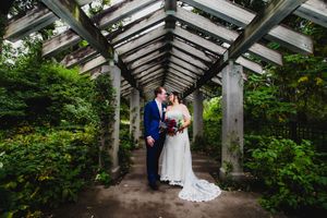 A couple in wedding attire shares a romantic moment under a wooden pergola surrounded by lush greenery.