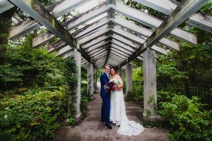 Wedding couple walking through an elegant garden pergola with white beams and green foliage.