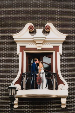 A couple shares a romantic moment on an ornate white balcony against a dark brick building facade.