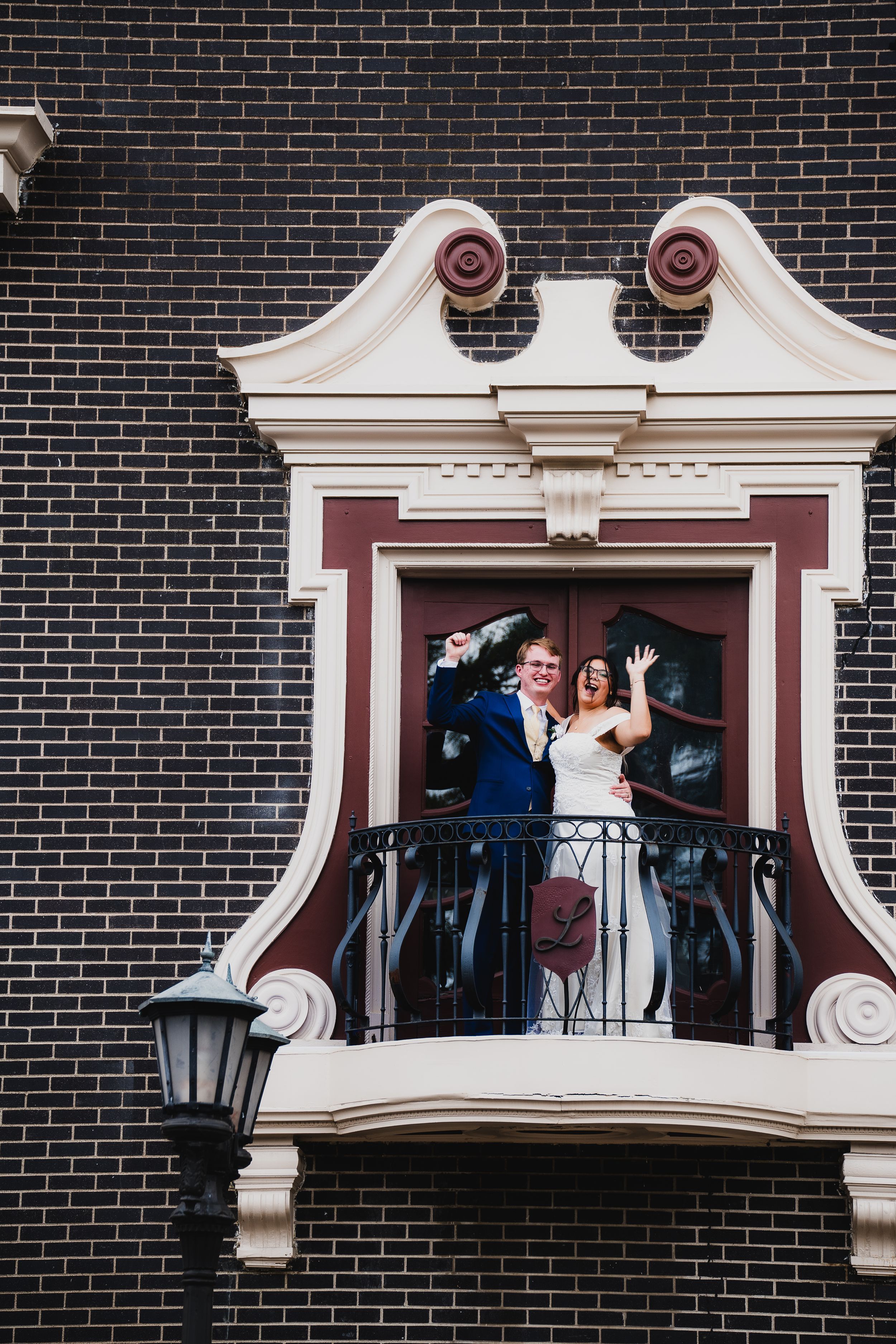 Wedding party poses on ornate balcony with white architectural details against dark brick wall.