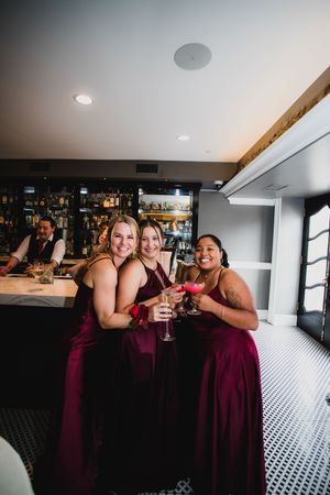 Group in burgundy dresses toasts with cocktails at modern bar with checkered floor.