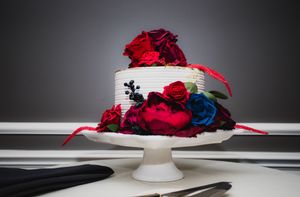 A vibrant red rose and blue flower arrangement sits elegantly displayed on a white cake stand against a dark background.