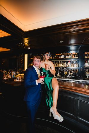 A couple shares an intimate moment at a dimly lit bar with a navy suit and emerald green dress.