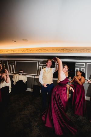 Wedding party members in burgundy dresses dance and celebrate at a reception.