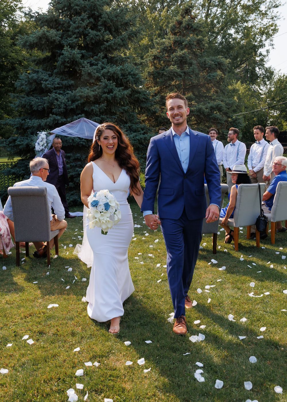 Couple walks through falling flower petals on a grassy lawn during an outdoor celebration.