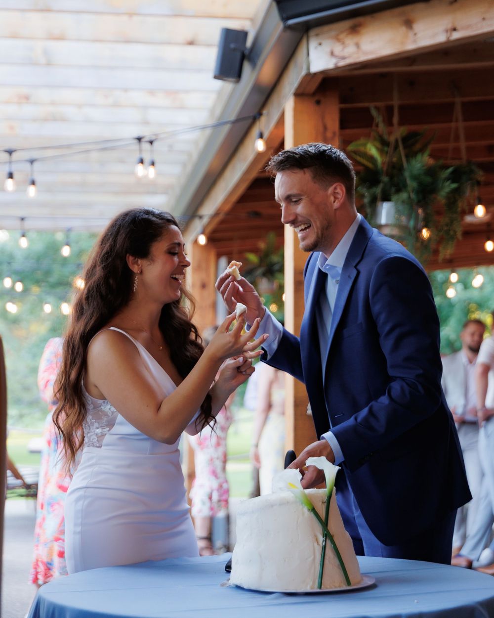 Couple playfully shares wedding cake during reception under covered outdoor venue with string lights.