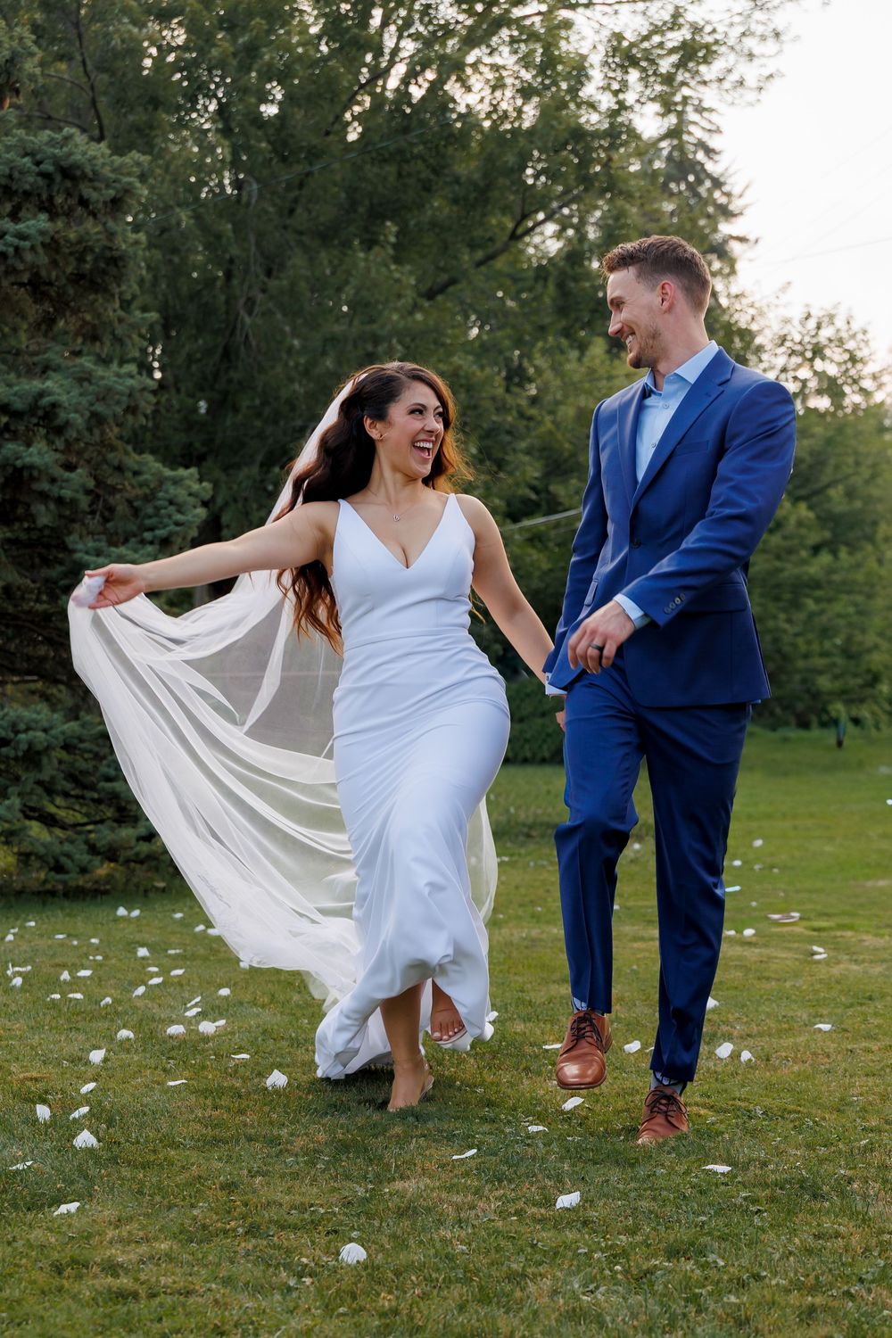 A couple in wedding attire playfully kicks fallen white petals on a grassy lawn during a sunny outdoor photoshoot.