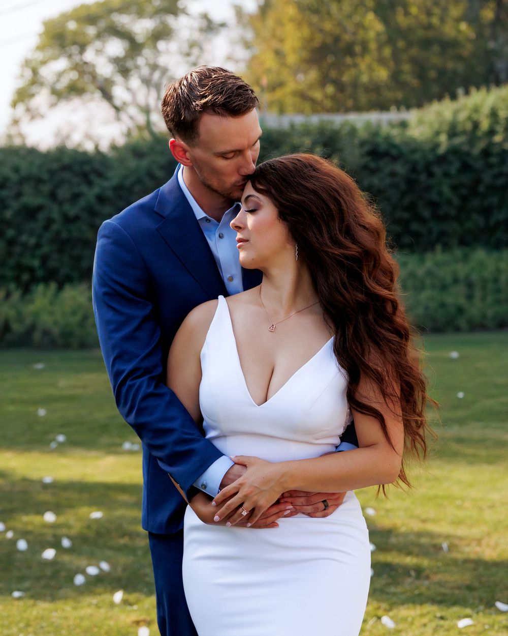 A couple embraces on a sunny lawn, with the bride wearing a white dress and groom in a navy blue suit.