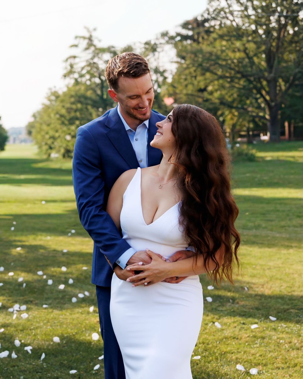 A couple in wedding attire embraces on a sunny lawn with scattered white flower petals around them.
