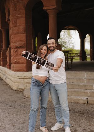 Smiling couple holding ultrasound photos during outdoor pregnancy announcement at historic brick building.