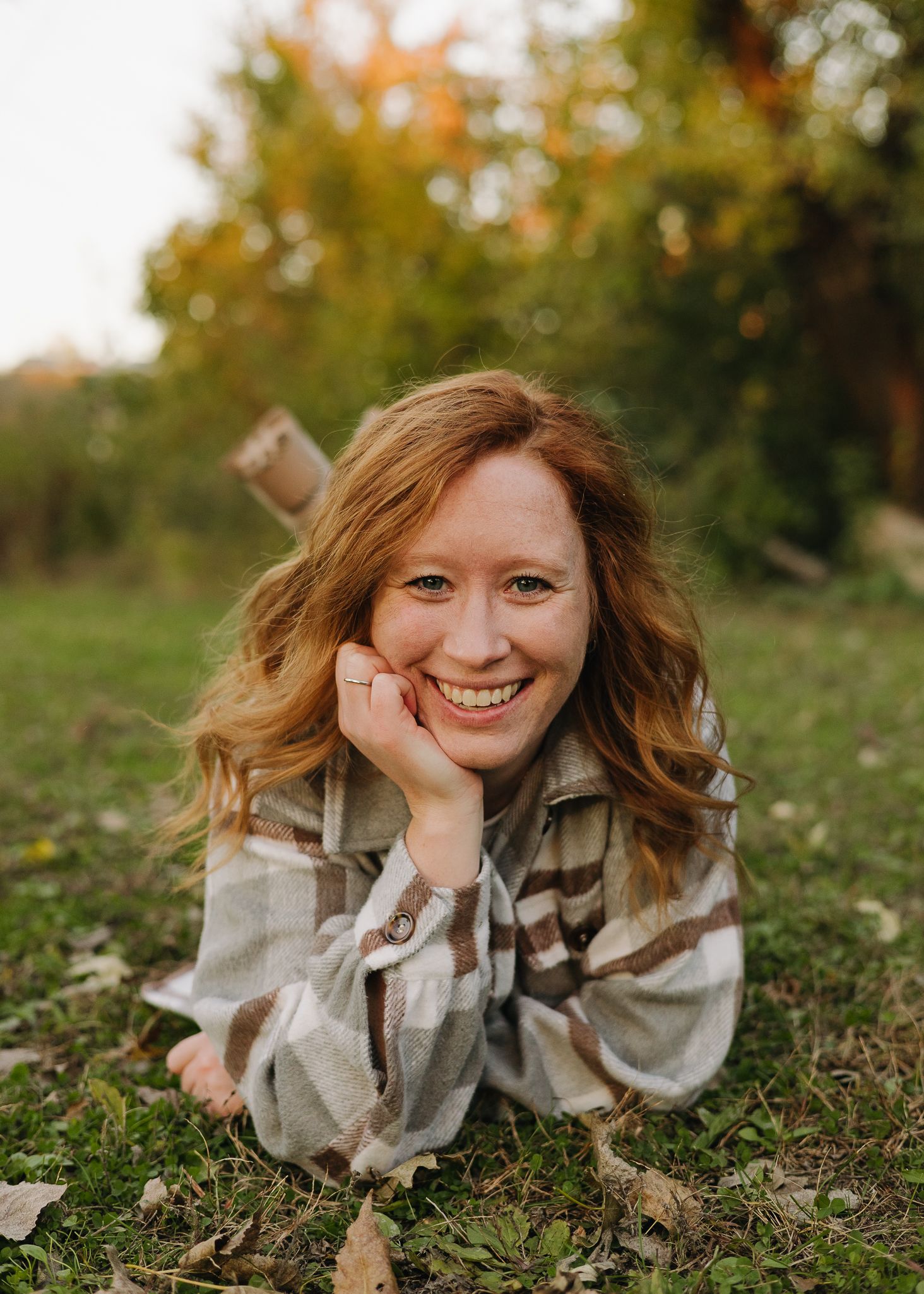 Smiling redhead woman lying on grass in autumn park wearing plaid flannel shirt.