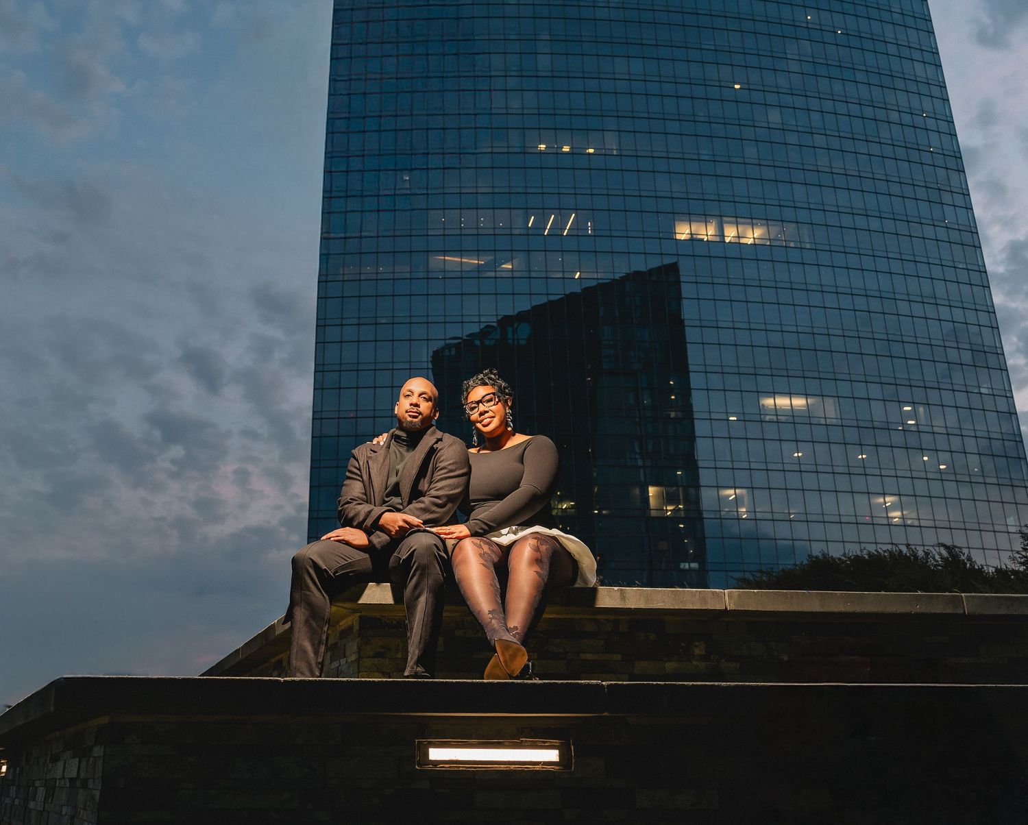 A couple wearing matching black and white hoodies smile at each other while sitting on a bench in a park setting.