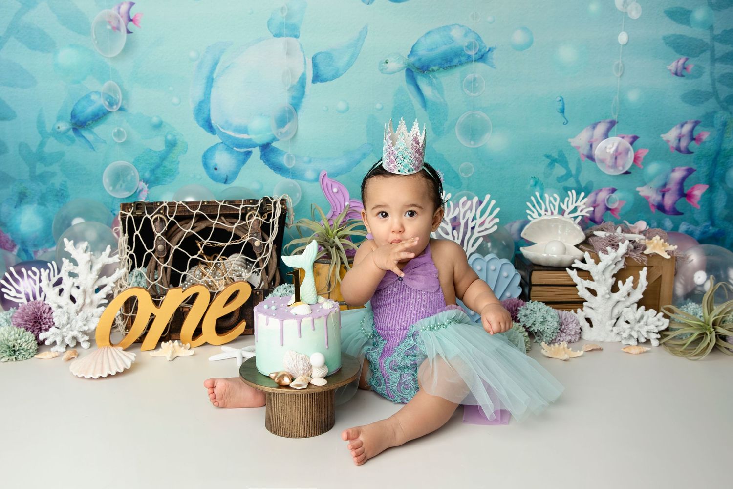 A baby in a turquoise tutu dress sits among floral decorations and party decorations for a first birthday celebration.
