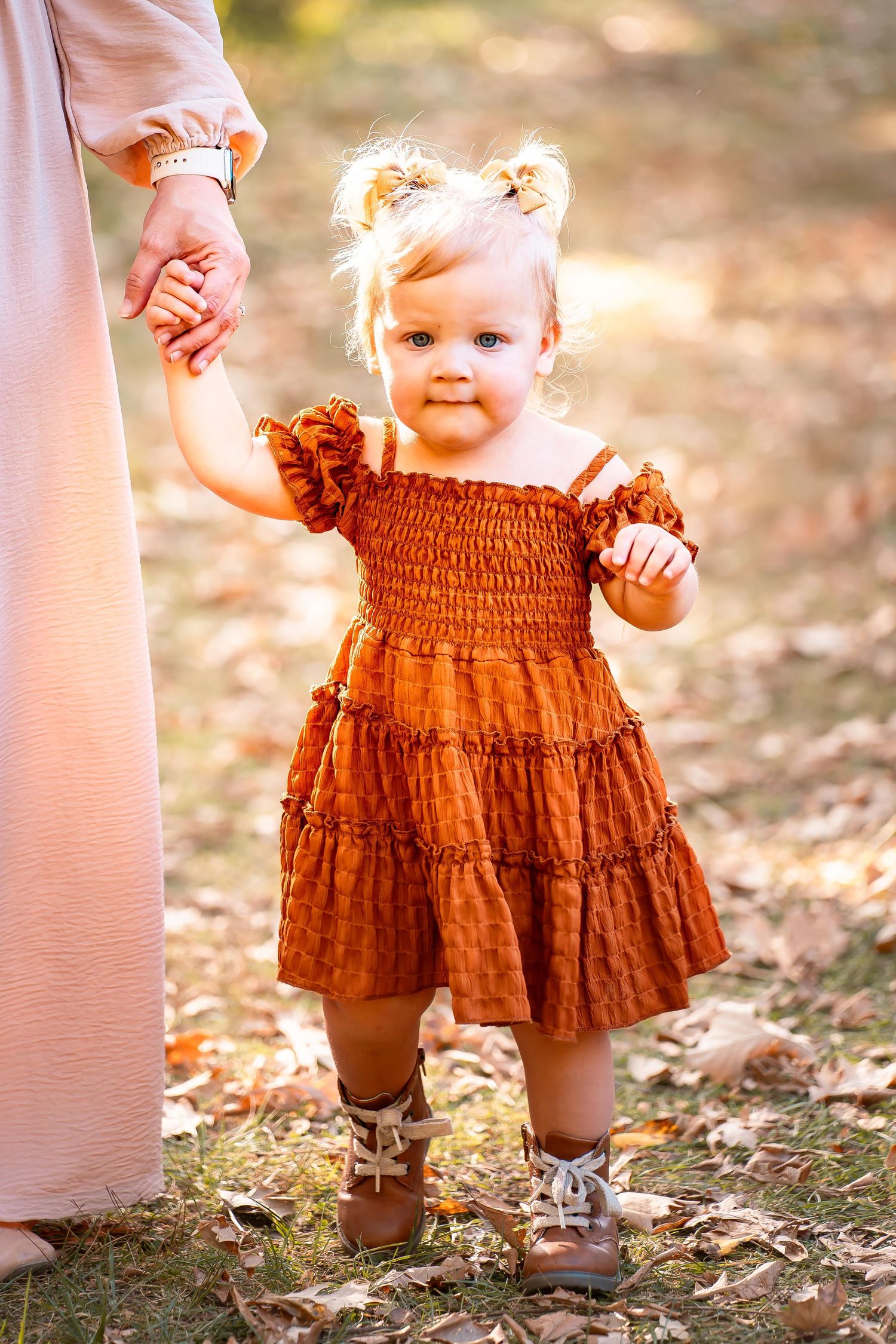 A toddler in a rust-colored smocked dress walks hand-in-hand with someone in autumn.
