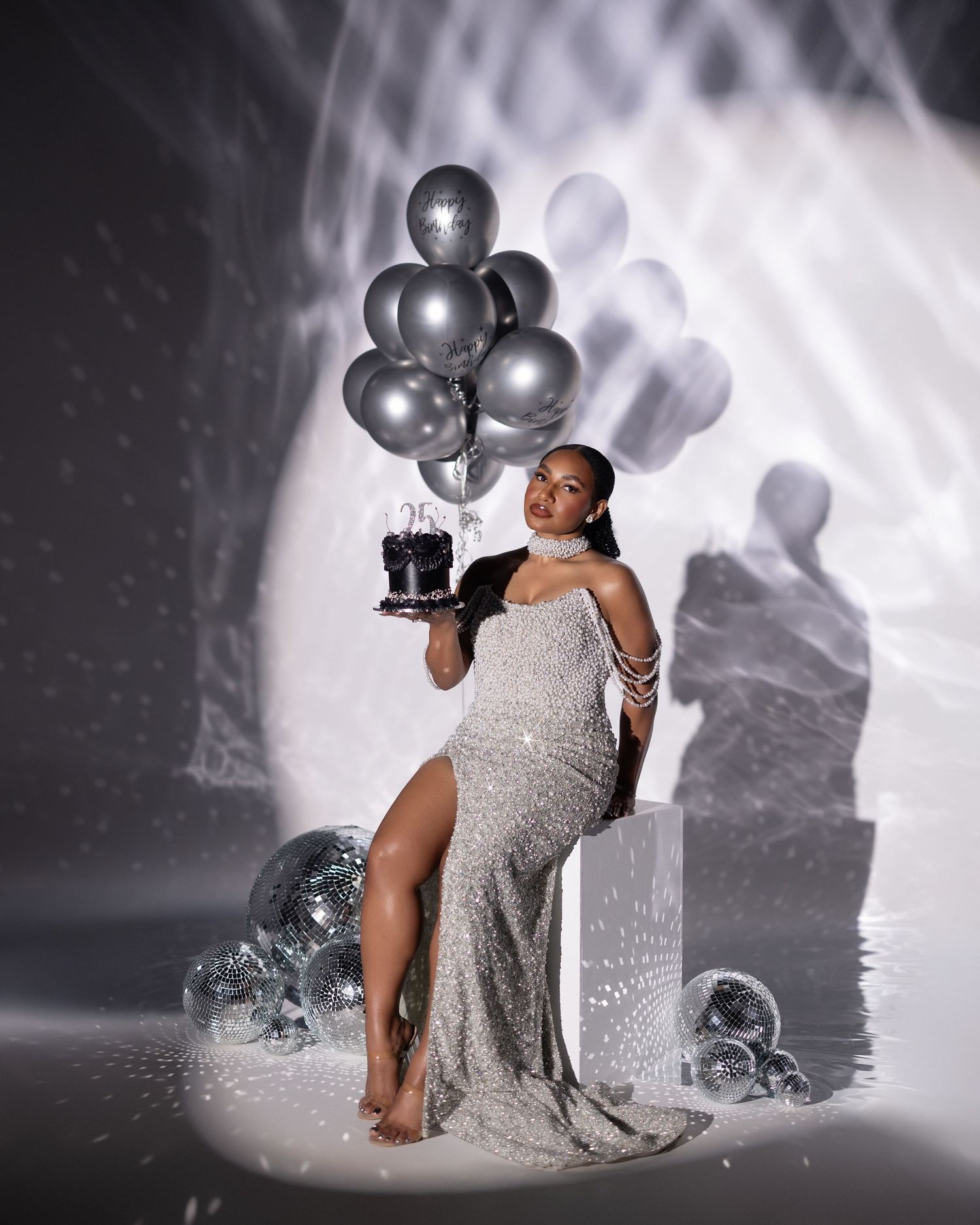 A figure in a sparkling silver gown poses elegantly with silver balloons against a dramatic monochrome backdrop.