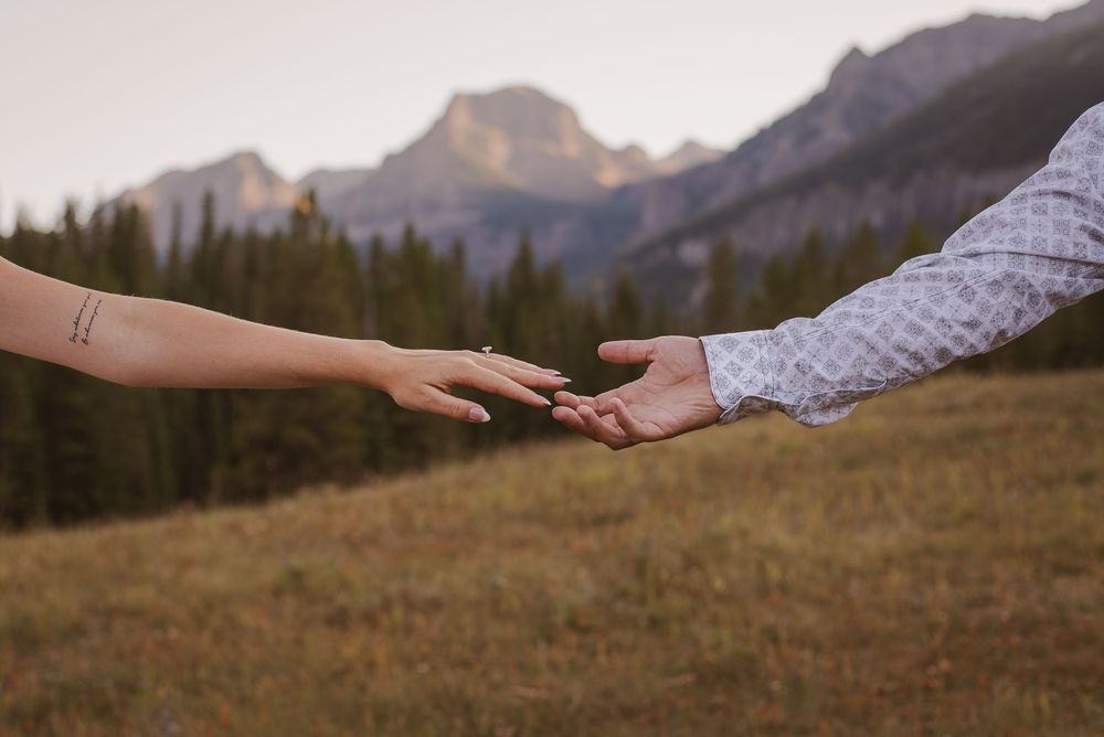 Reaching hands connect across a mountain meadow with dramatic peaks in the background.