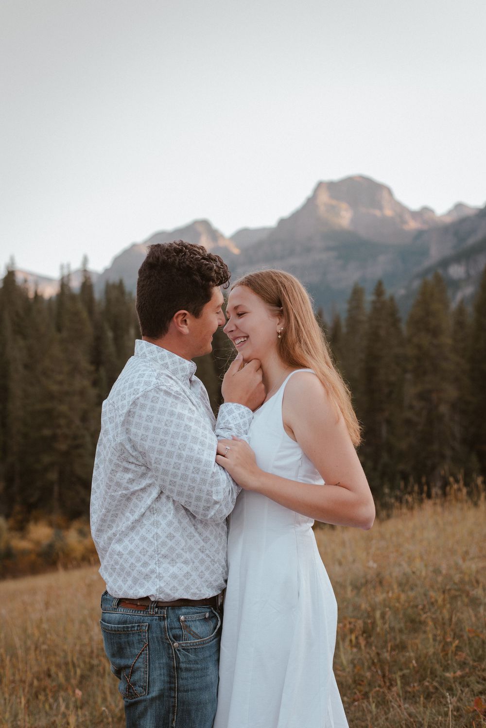 A couple shares tender moments in a mountain meadow with evergreen trees and peaks in the background.