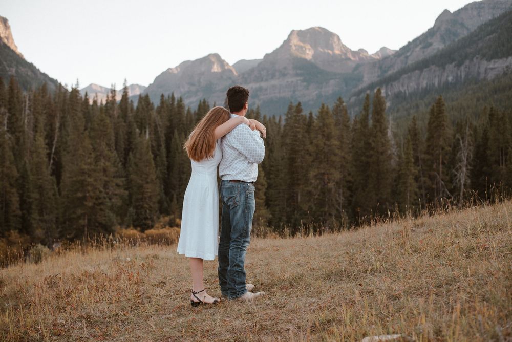 A couple walks hand in hand through a mountain meadow with dramatic peaks in the distance.