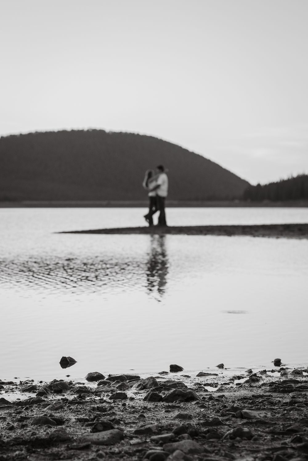 A silhouetted couple embraces by a lake with hills in the background.